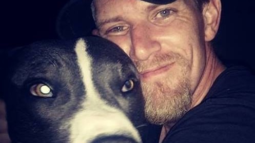 A man wearing a black t-shirt and baseball caps hugs his dog