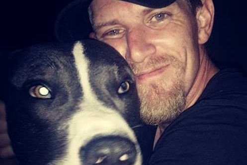A man wearing a black t-shirt and baseball caps hugs his dog