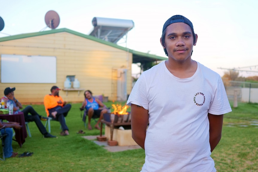 A young Aboriginal man in a T-shirt and cap smiling with his family sitting around a fire in the background.