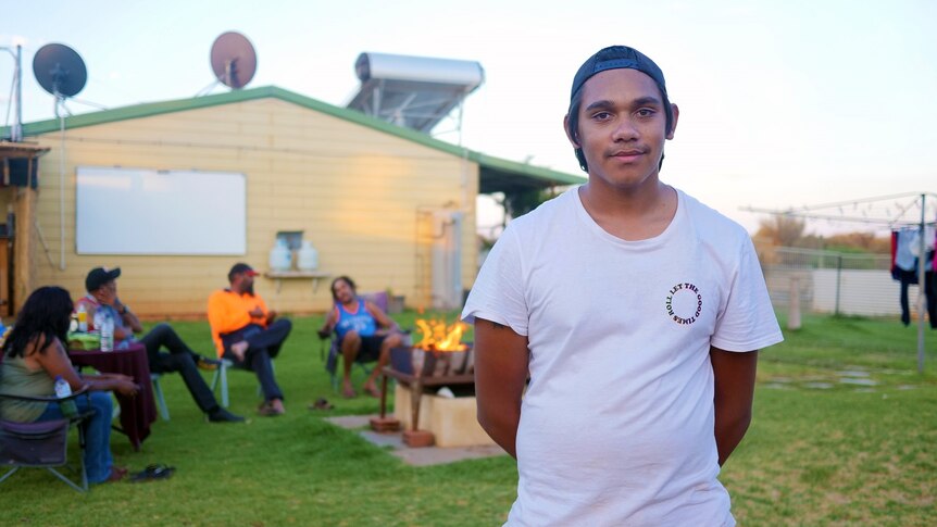 A young aboriginal man in a tshirt and cap smiling with his family sitting around a fire in the background