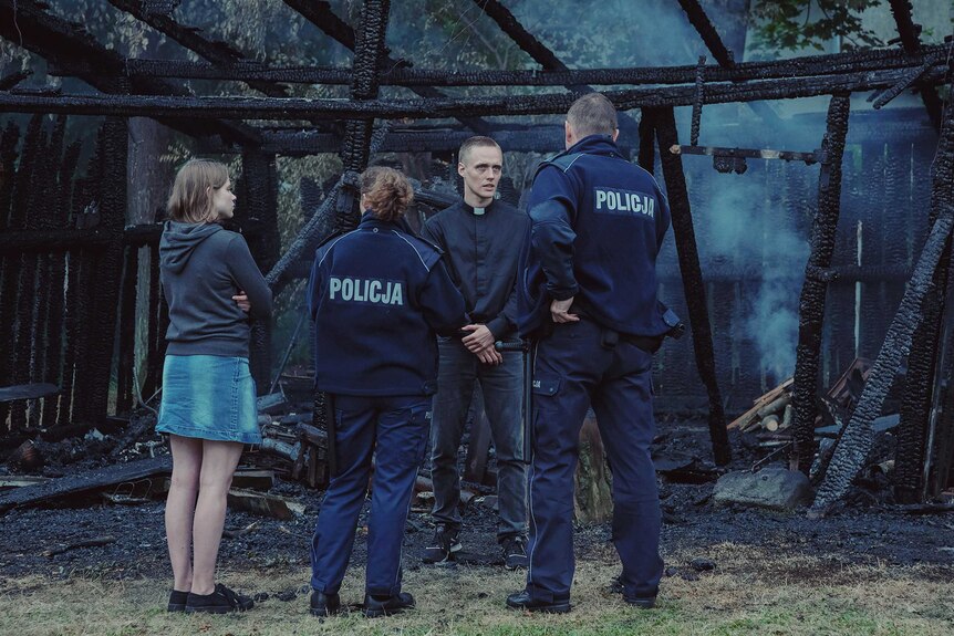 A scene from the film Corpus Christi with a young priest speaking to police in front of a burnt building