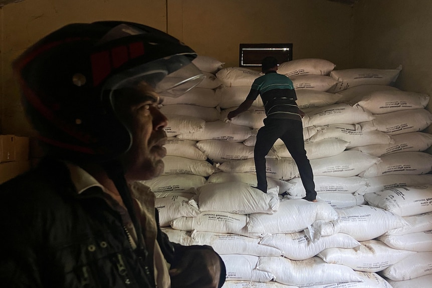 A man in a helmet looks on from the foreground as another man climbs over stacks of white fertiliser bags in a warehouse.