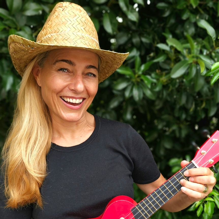Natacha Curnow, wearing a straw hat and holding a bright pink ukulele, standing in front of a lush green hedge.
