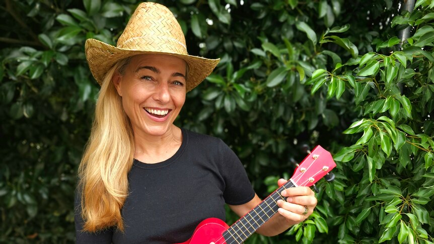 Natacha Curnow, wearing a straw hat and holding a bright pink ukulele, standing in front of a lush green hedge.