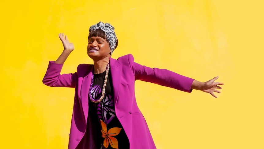 Against a bright yellow backdrop, a woman in a purple blazer, cheque headscarf, and Pacific-style dress poses.