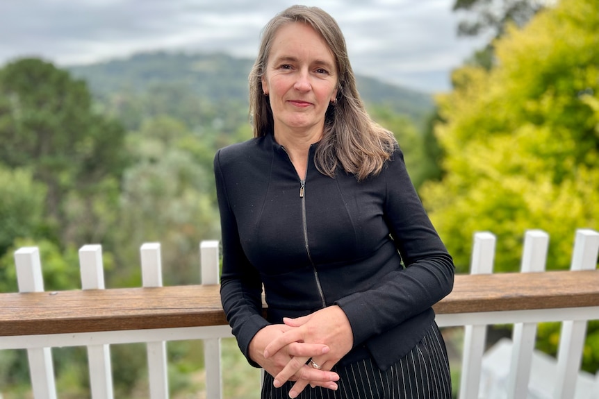 A woman wearing black stands on a balcony with a green landscape behind her.