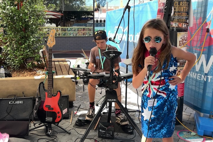 a boy on key board, a boy on drums and a girl singing, wearing an australian flag theme dress and sunglasses busk in Tamworth