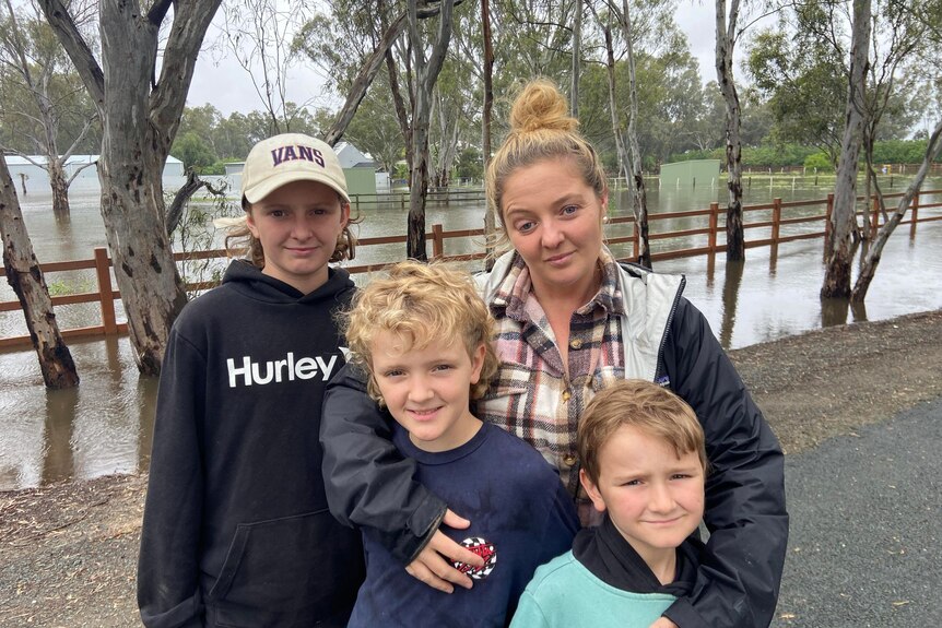 A sad looking woman with blone hair tied up in a bun, stands with three boys in front of flood water.