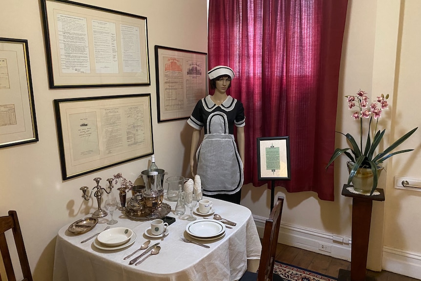 A museum exhibit of an old room in a hotel, including a mannequin standing next to a set table. 