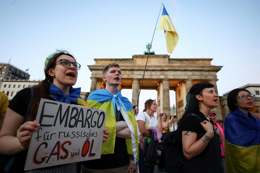 Protesters hold signs and wave yellow and blue flags in front of the Brandenburg Gate.