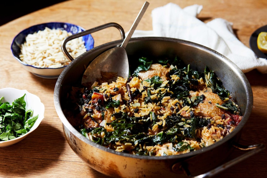 A deep fry pan with chicken thighs, risoni and silver beet with chopped almonds and parsley in bowls as toppings, family dinner.