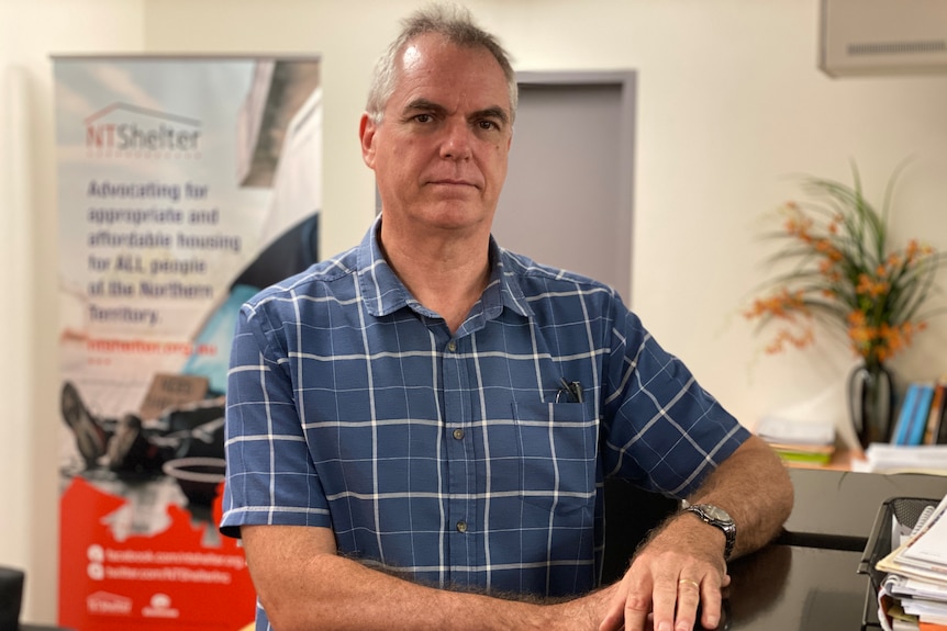 A man in a collared shirt standing in an office, looking serious.