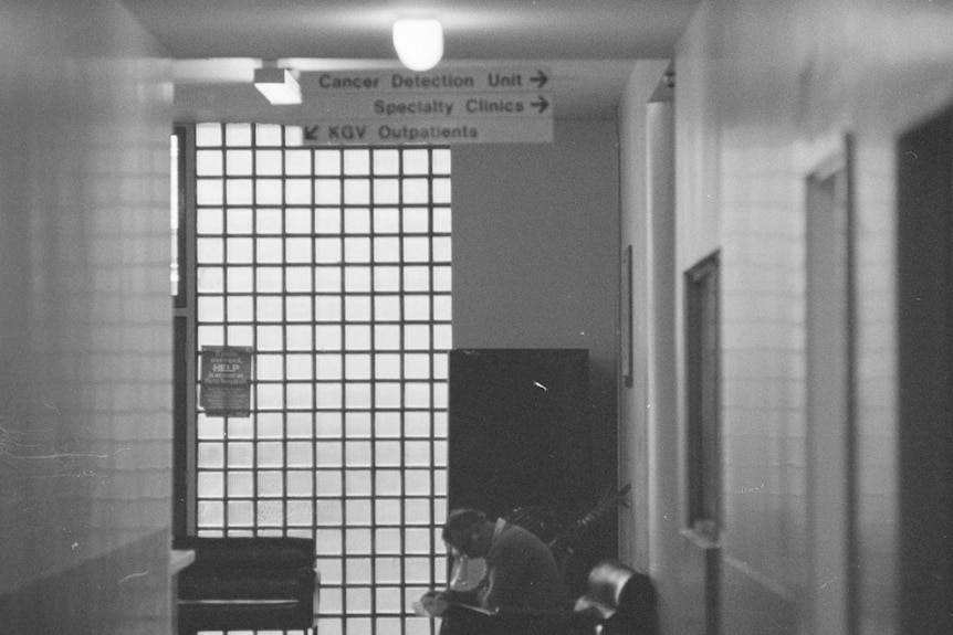 A man looks down at his hands as he sits in a chair in an otherwise empty hospital corridor.