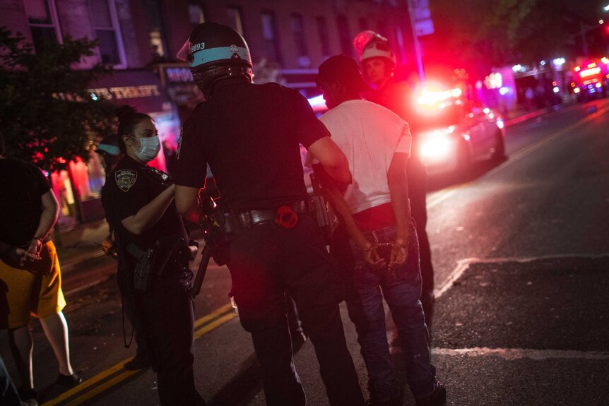 At night, lit by the red and blue flashes of police sirens, two police arrest a black protester.