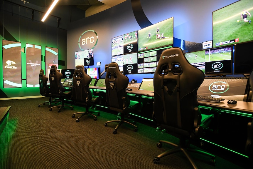 A picture shows a room with a series of desks with monitors and big screens showing footage from an AFL match.