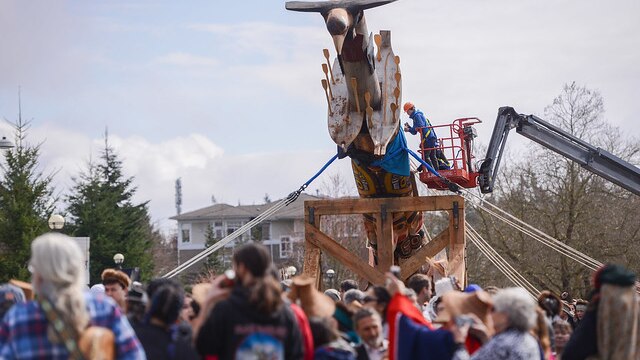 A crowd of people stand around while a crane lifts a traditional pole into place.