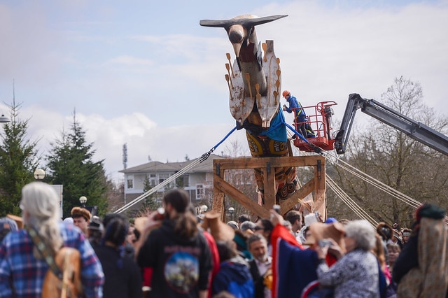 A crowd of people stand around while a crane lifts a traditional pole into place.