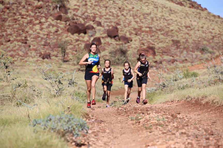 A woman runs through red dust and dry grass alongside three young girls.