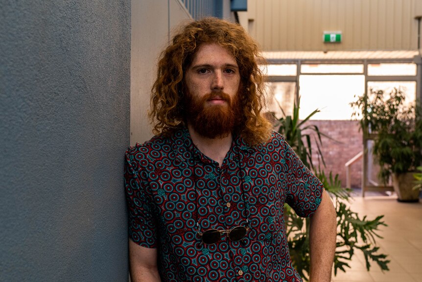 A young man with red hair and a beard and sunglasses hanging from his neck has a steely and a serious look.