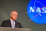 Scott Morrison speaks behind a silver metal lectern beside national flags as a Nasa logo is projected onto a wall behind him.