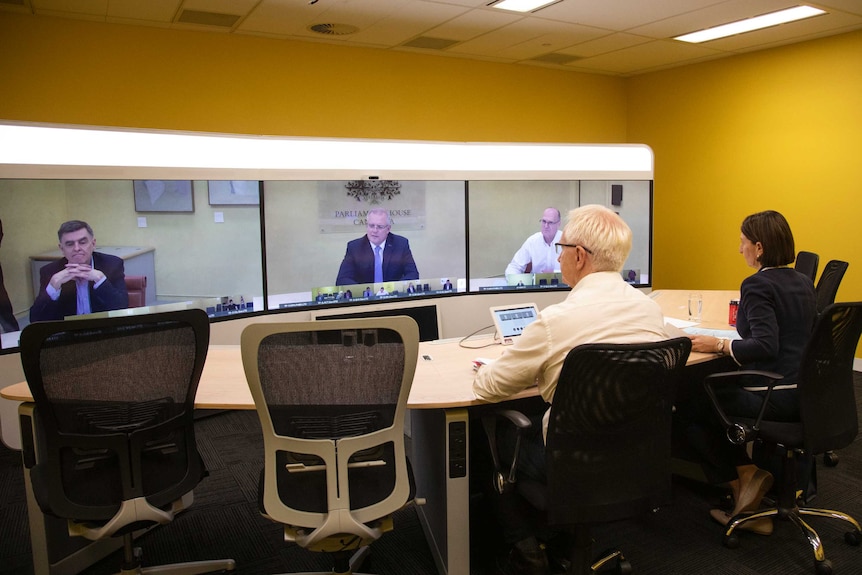 Gladys Berejiklian and Tim Reardon sit at a desk and watch while Scott Morrison speaks on a screen.