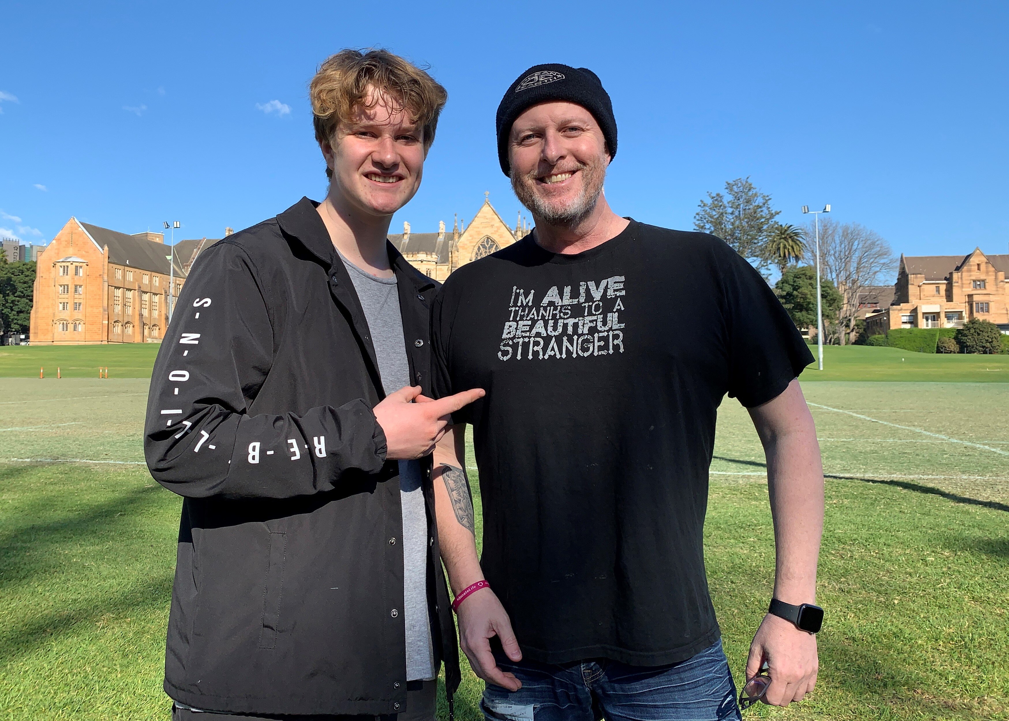 Two men standing outside smiling. The younger one pointing to the older one.