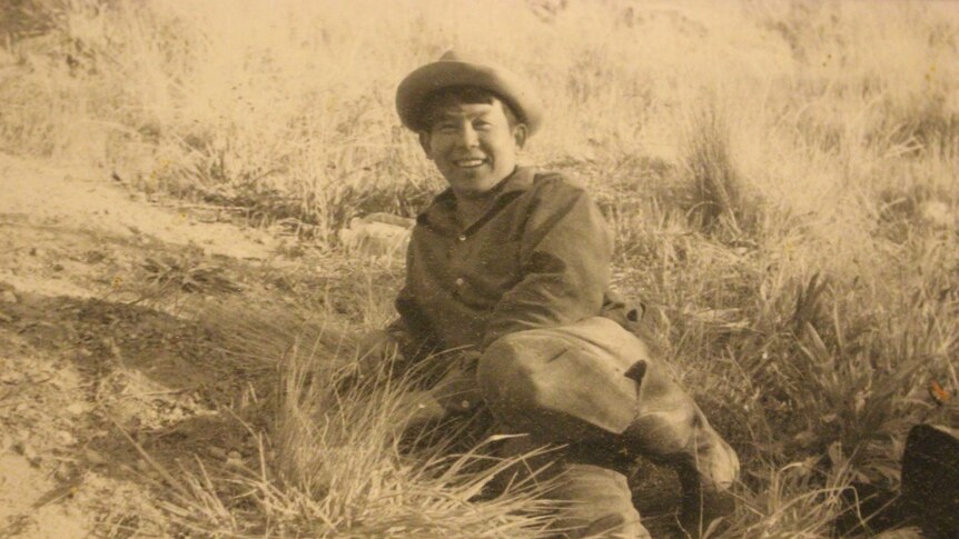 A sepia, old photo shows a man smiling, lying in a grassy field.