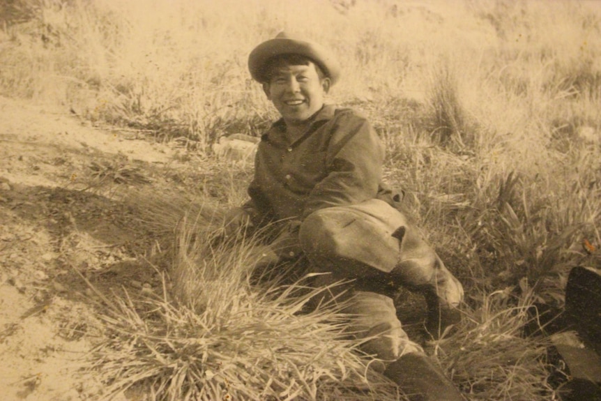 A sepia, old photo shows a man smiling, lying in a grassy field.