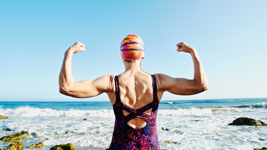 The back of a woman standing, facing the ocean, wearing a swimsuit and swim cap, and flexing the muscles of both raised arms.
