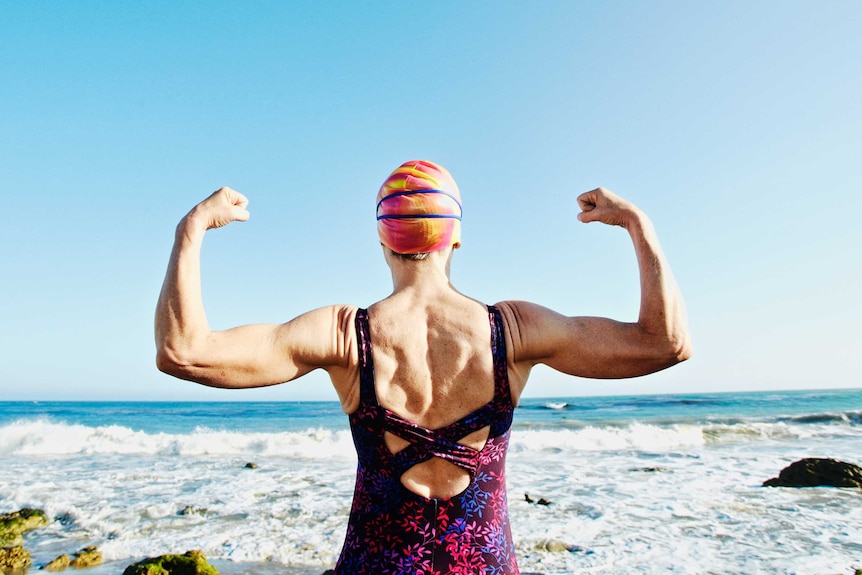 The back of a woman standing, facing the ocean, wearing a swimsuit and swim cap, and flexing the muscles of both raised arms.