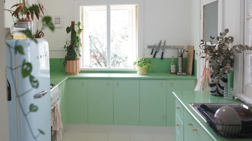 A kitchen is seen with a blue fridge to the left, green countertops and cupboards in front. The floor is white