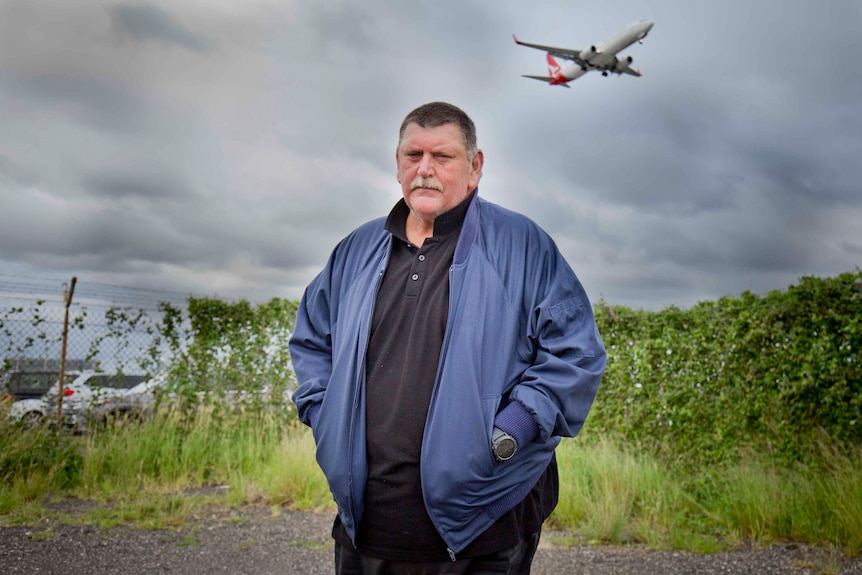 A portrait of Bob Pickard looking seriously at the camera, as a plane fli