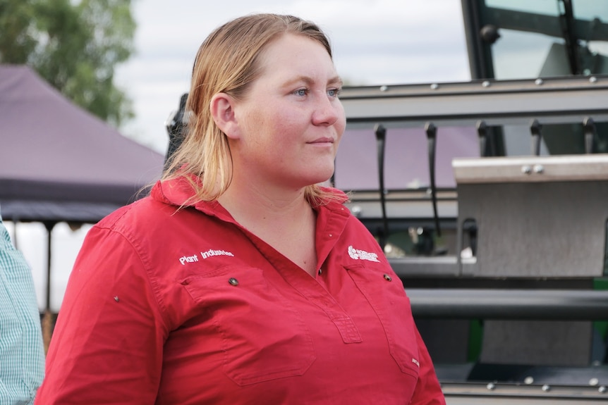 A young woman in a bright coloured workshirt watches on standing in front of a large piece of harvesting equipment on a farm