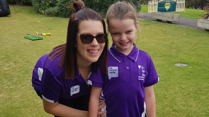 Katherine and Ella stand next to each other on an oval, wearing purple shirts. Katherine is bending down to Ella's height.