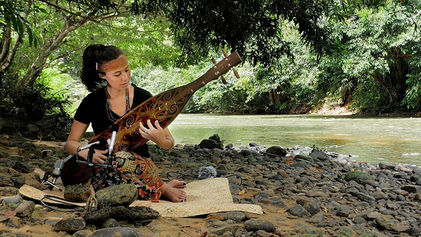 A woman sits on a rug by a river in the jungle playing a wooden stringed instrument (a Sape')