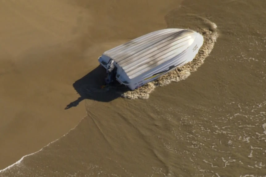 A tinnie lies overturned on the sand with water washing up against it.
