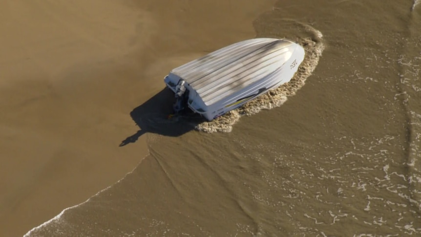 A tinnie lies overturned on the sand with water washing up against it.