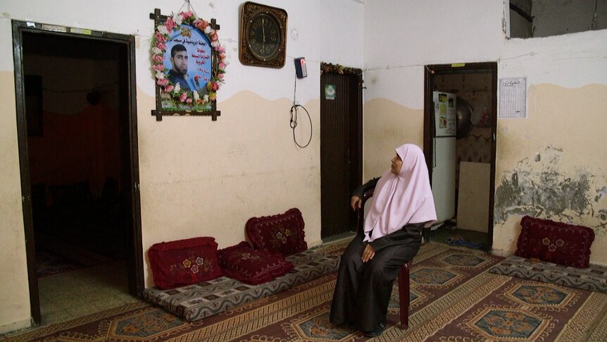A woman named Sabah sits in a bare house looking at a photo of her son on the wall.