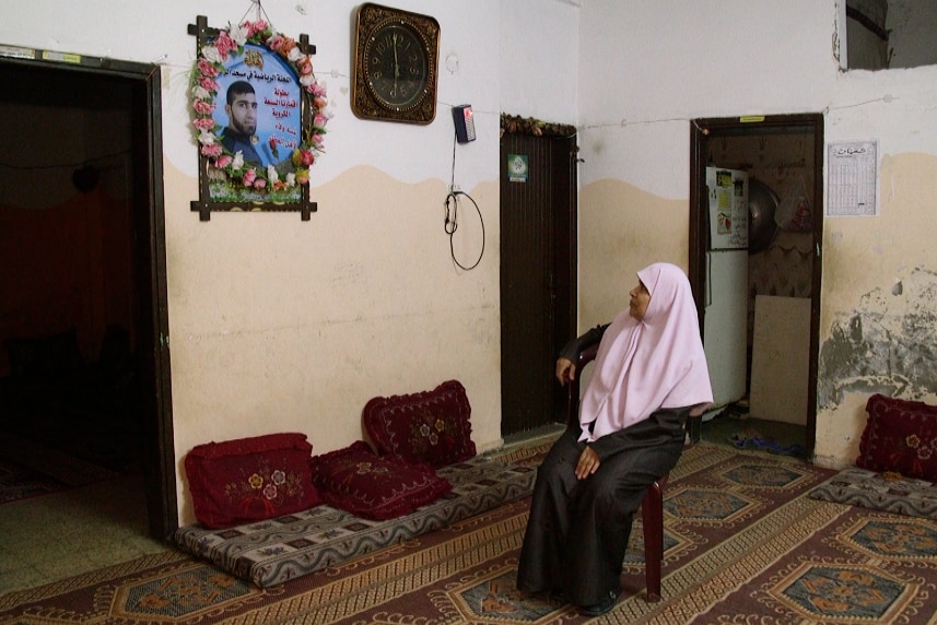 A woman named Sabah sits in a bare house looking at a photo of her son on the wall.