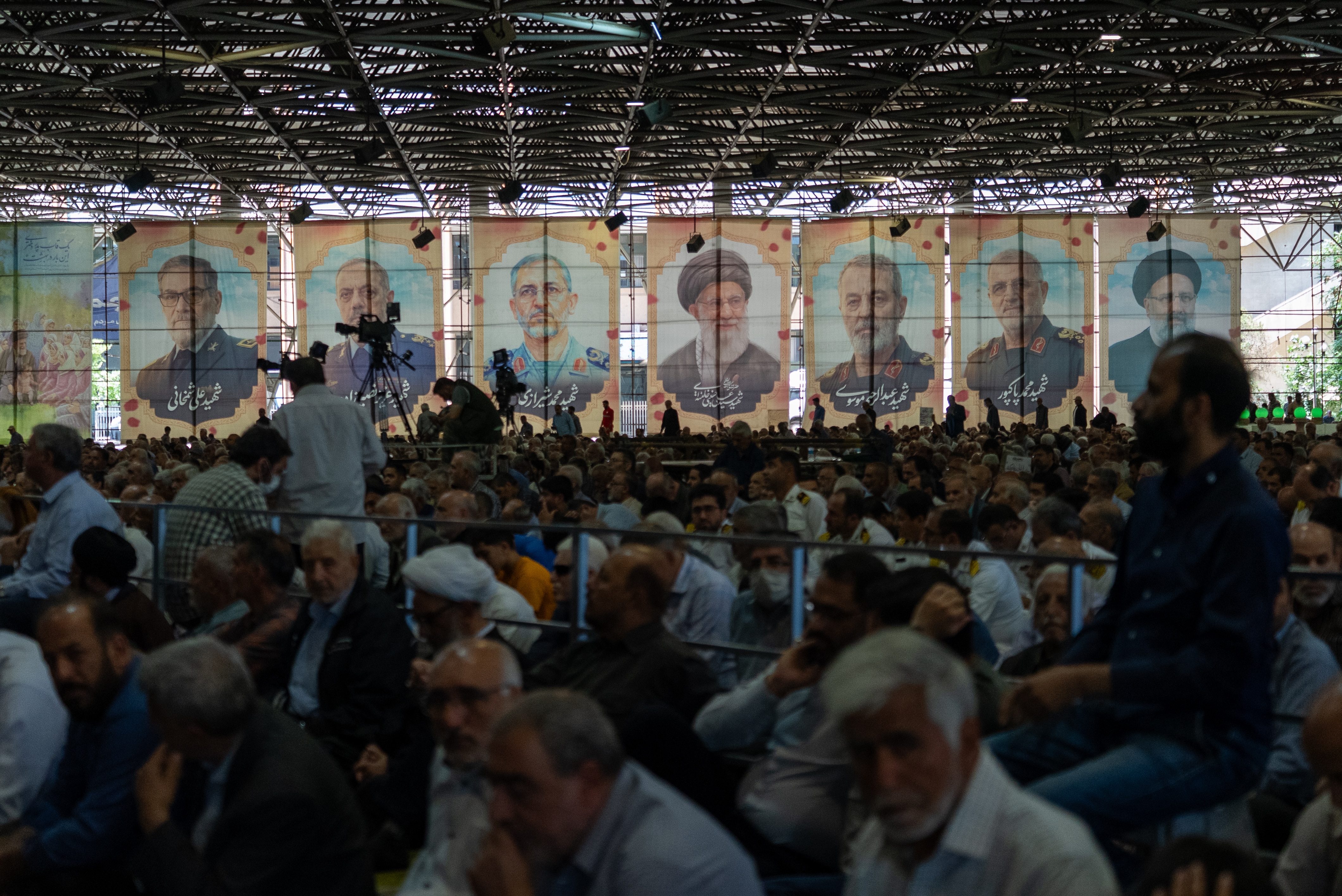 A large crowd of men and boys sit in a hall, in front of large photos of religious and military leaders.
