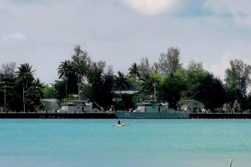 A person kayaks past a larger vessel moored on calm, turquios waters at a long jetty with palm tress behind.