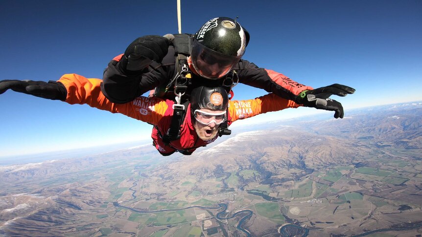 A man with a blonde beard skydiving in tandem with another man, his arms outstretched