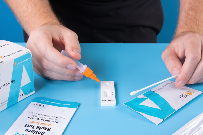 A close-up of a person using a rapid antigen test, holding a swab in one hand and a dropper bottle over the test in the other.