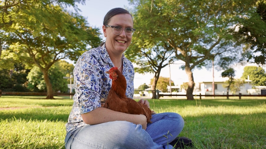A smiling woman sits on some grass, cradling a chicken.