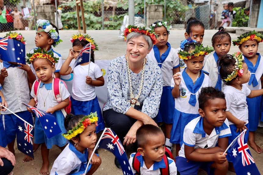 Penny Wong, wearing a floral wreath in her hair, kneels next to Kiribati children holding Australian flags