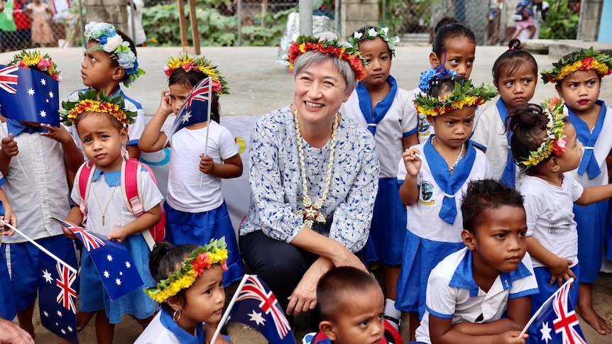 Penny Wong, wearing a floral wreath in her hair, kneels next to Kiribati children holding Australian flags