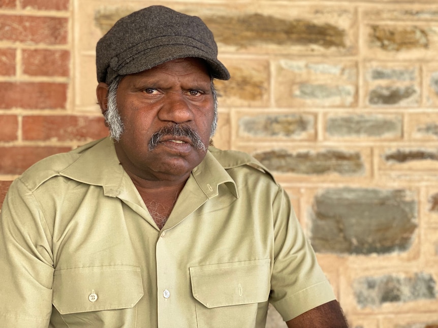 A man in a cap and tan shirt looks stern in front of an old brick wall