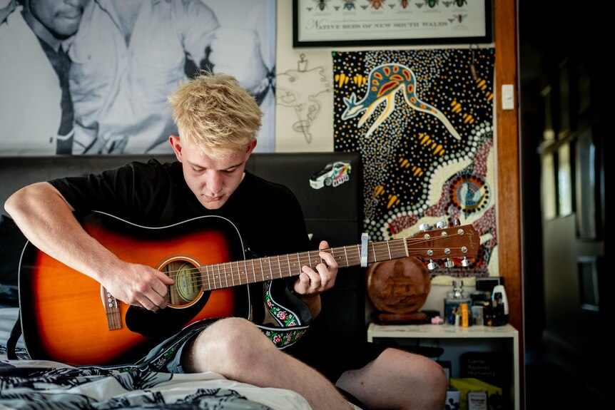 Boy sits on his bed while strumming an acoustic guitar