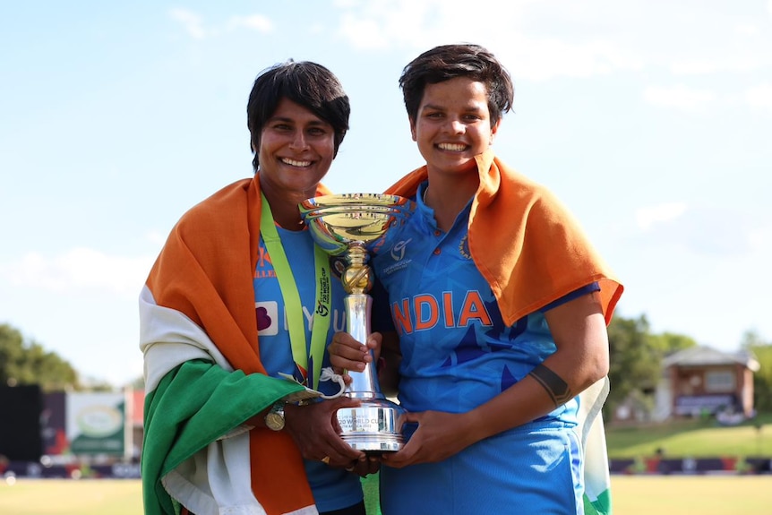 Nooshin Al Khadeer and Shafali Verma hold a trophy and smile while wearing Indian jerseys.