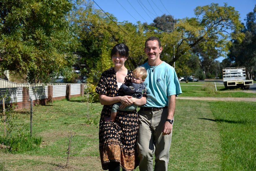 A young couple poses for a photo in the sun with their baby, on a grassy lawn